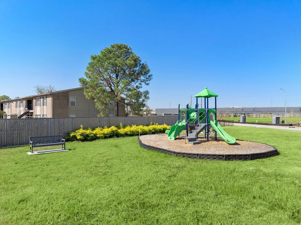 A small play structure in a grassy area with a tree and a building in the background.